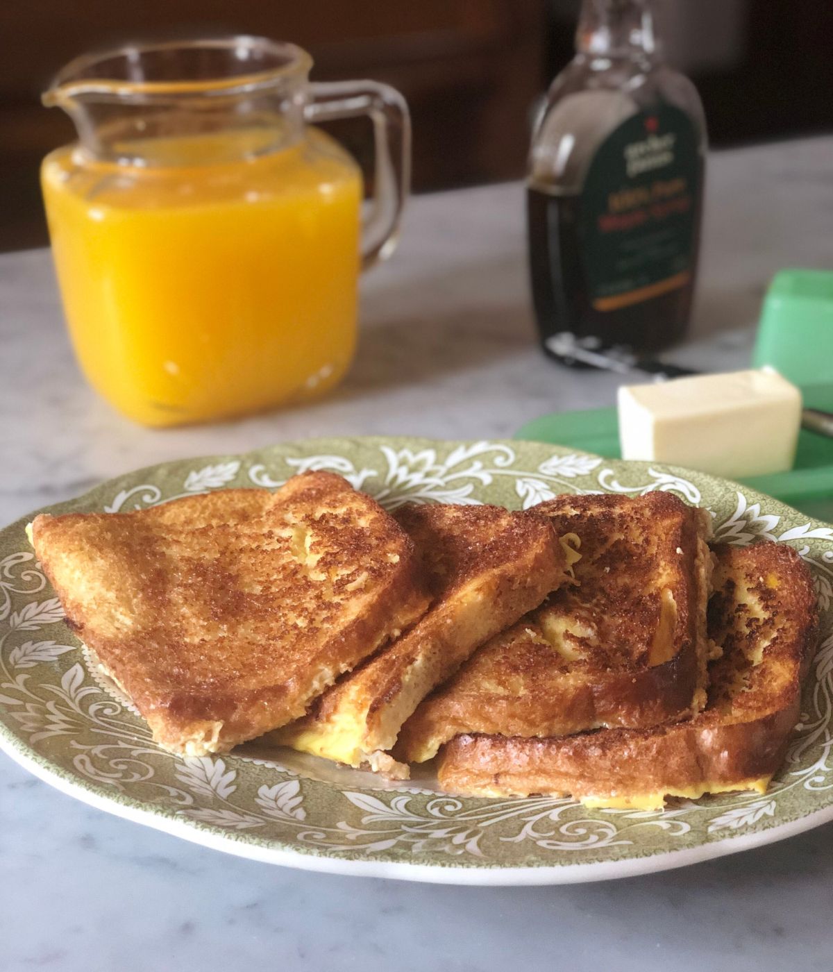four slices of baked french toast on a plate with orange juice, syrup and butter in the background